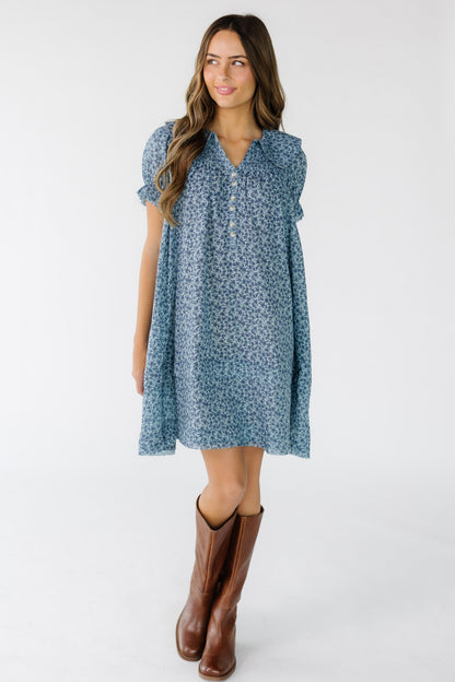 Young woman wearing a modest blue floral print short dress with short puff sleeves, rounded collar, short button placket and smocked front and back yoke.