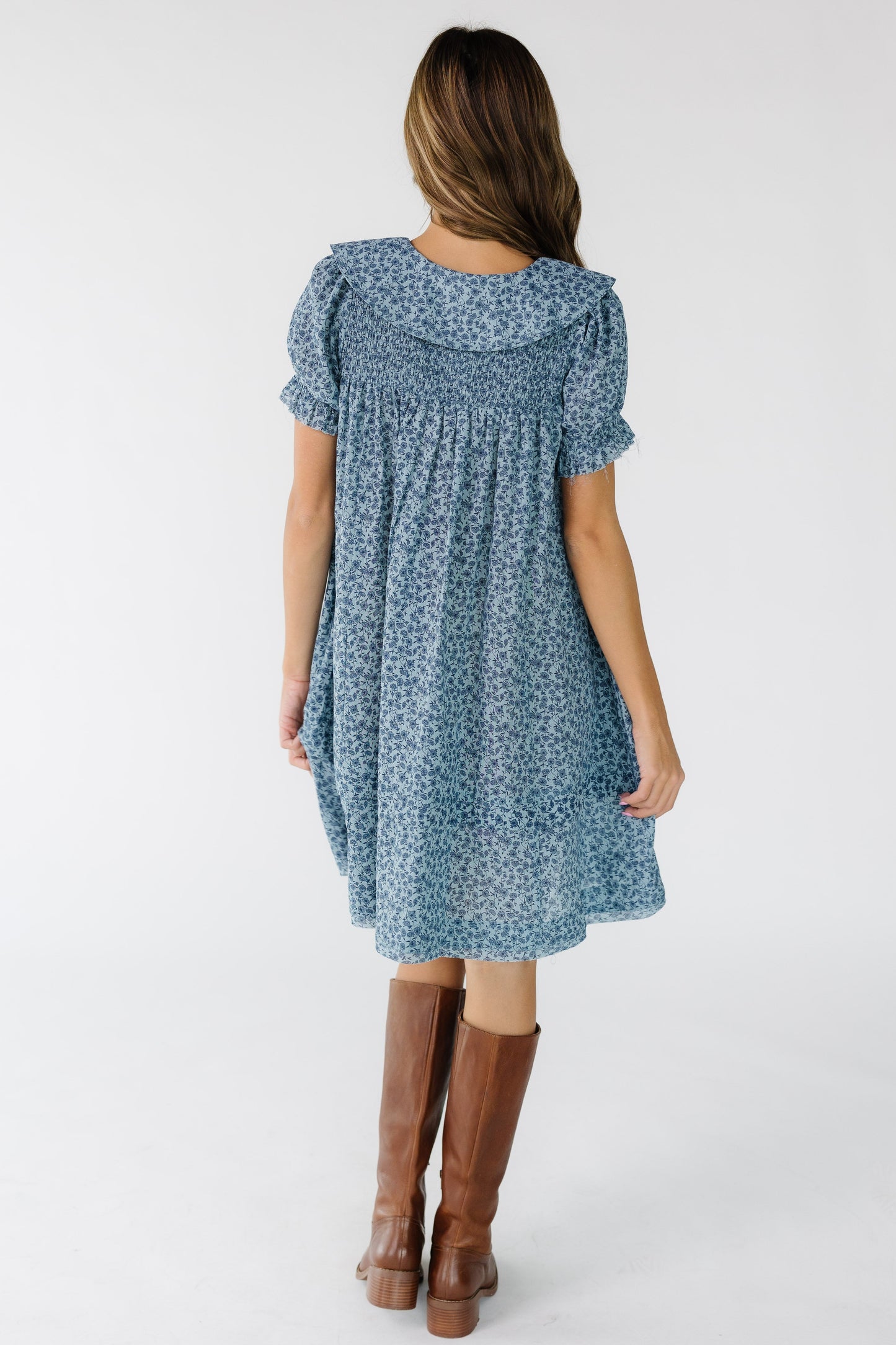 Back view of a young woman wearing a blue floral print, loose fitting dress with a smocked back yoke and wide collar.
