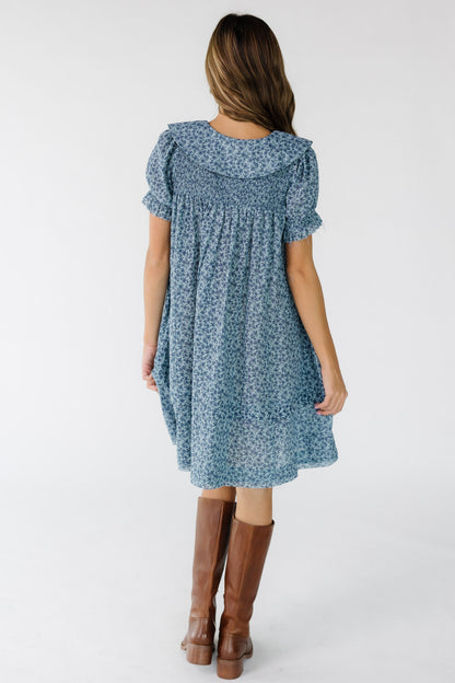 Back view of a young woman wearing a blue floral print, loose fitting dress with a smocked back yoke and wide collar.