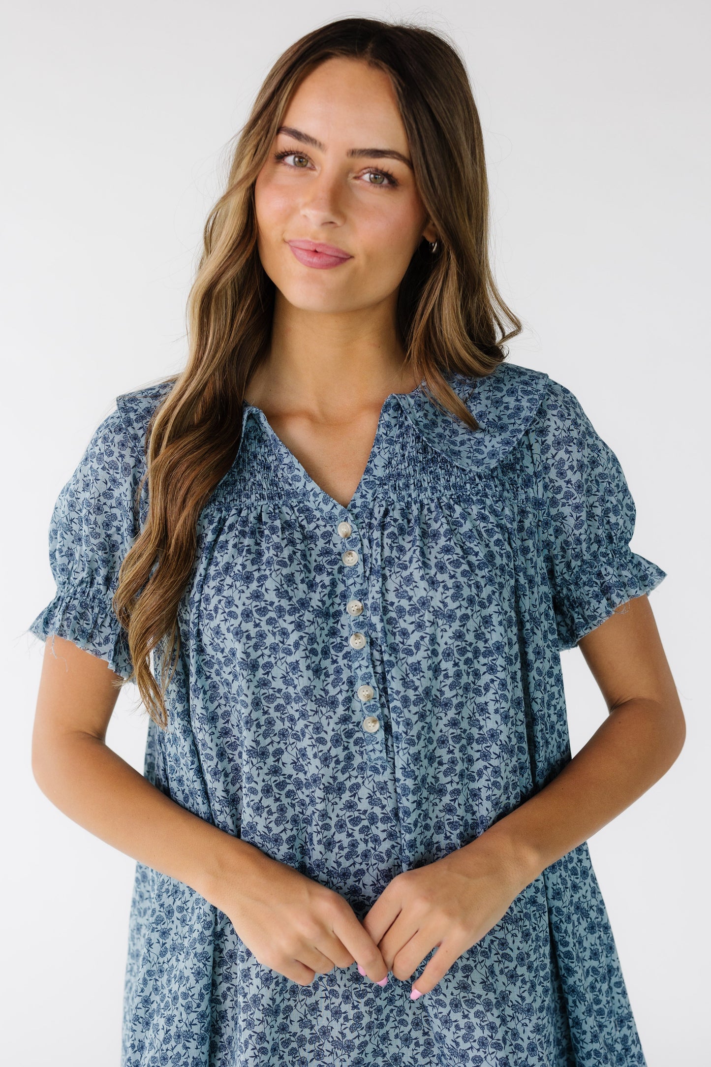 Close view of a young woman wearing a blue floral print dress with a short button placket, V-neckline and front and back smocked yoke.