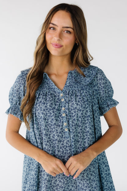 Close view of a young woman wearing a blue floral print dress with a short button placket, V-neckline and front and back smocked yoke.