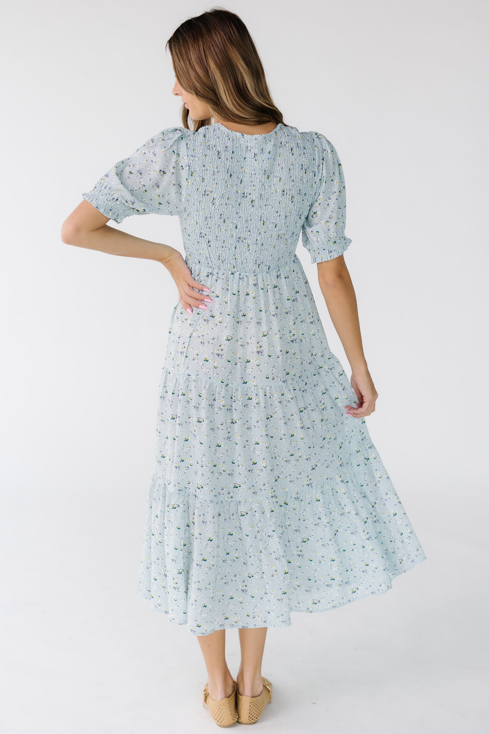 Back view of a young woman wearing a light blue floral print midi dress with multiple gathered tiers and a smocked back bodice.
