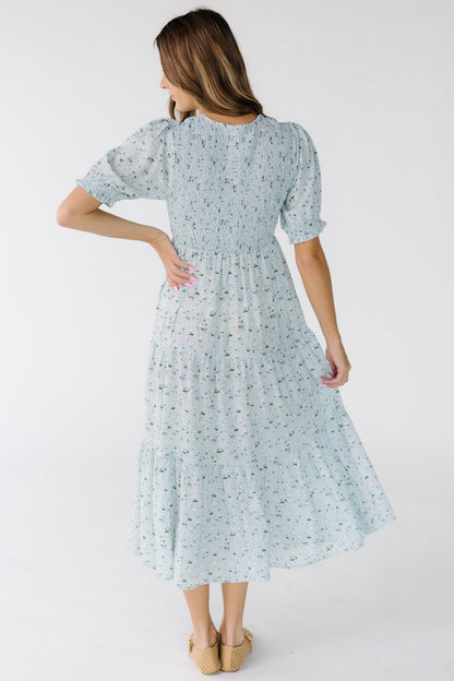 Back view of a young woman wearing a light blue floral print midi dress with multiple gathered tiers and a smocked back bodice.