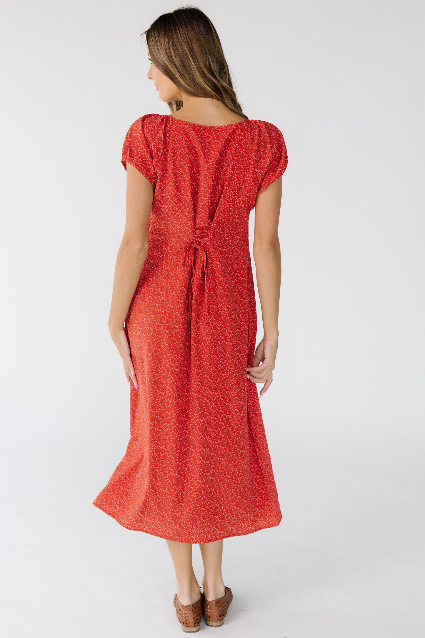 Back view of a woman wearing a red floral print column midi dress that laces at the center back with short puff sleeves.