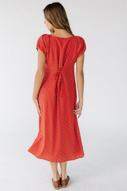Back view of a woman wearing a red floral print column midi dress that laces at the center back with short puff sleeves.