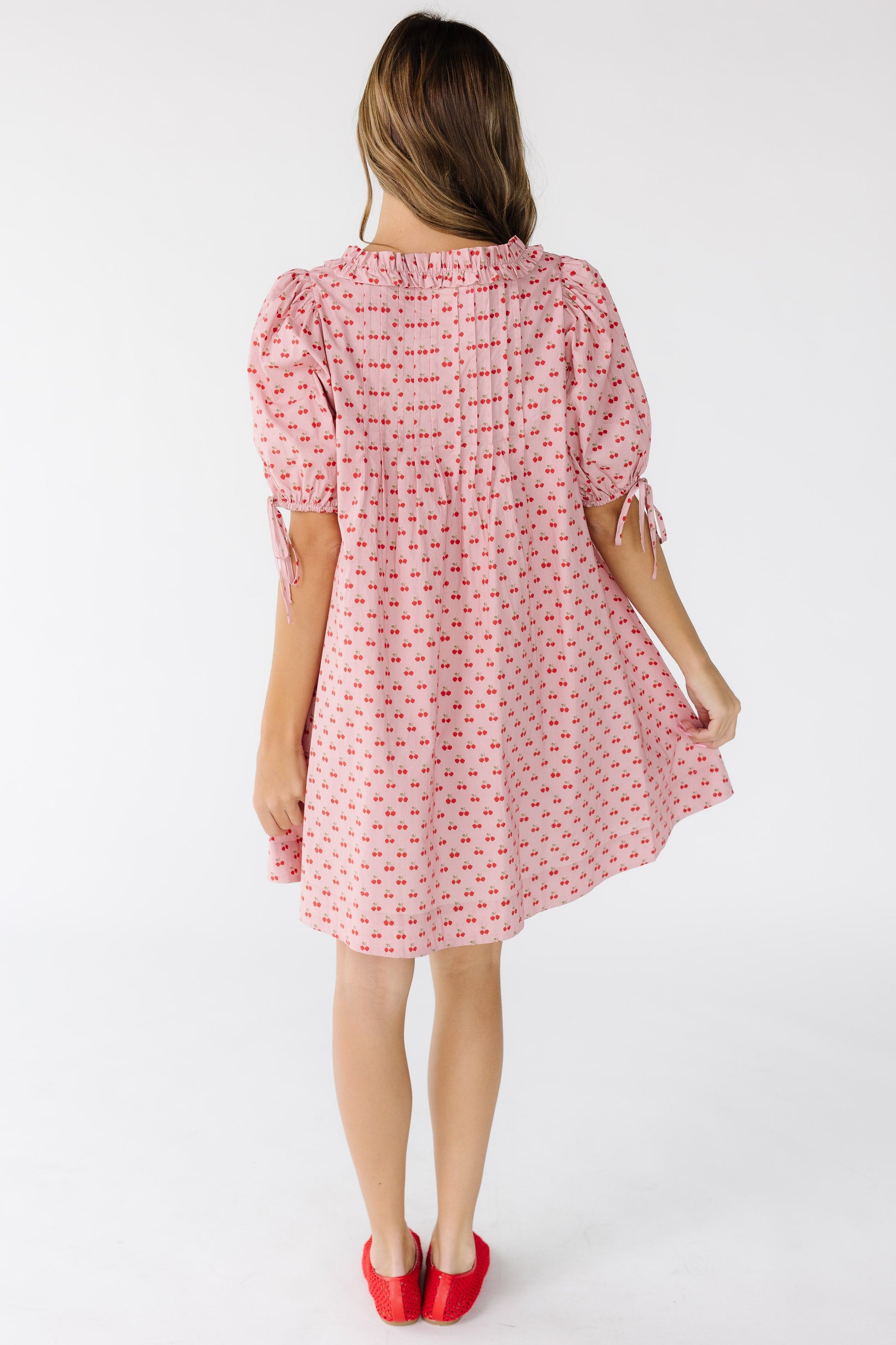 Back view of a young woman wearing a pink cherry print loose fitting short dress with back sewn in pleats and large balloon sleeves.