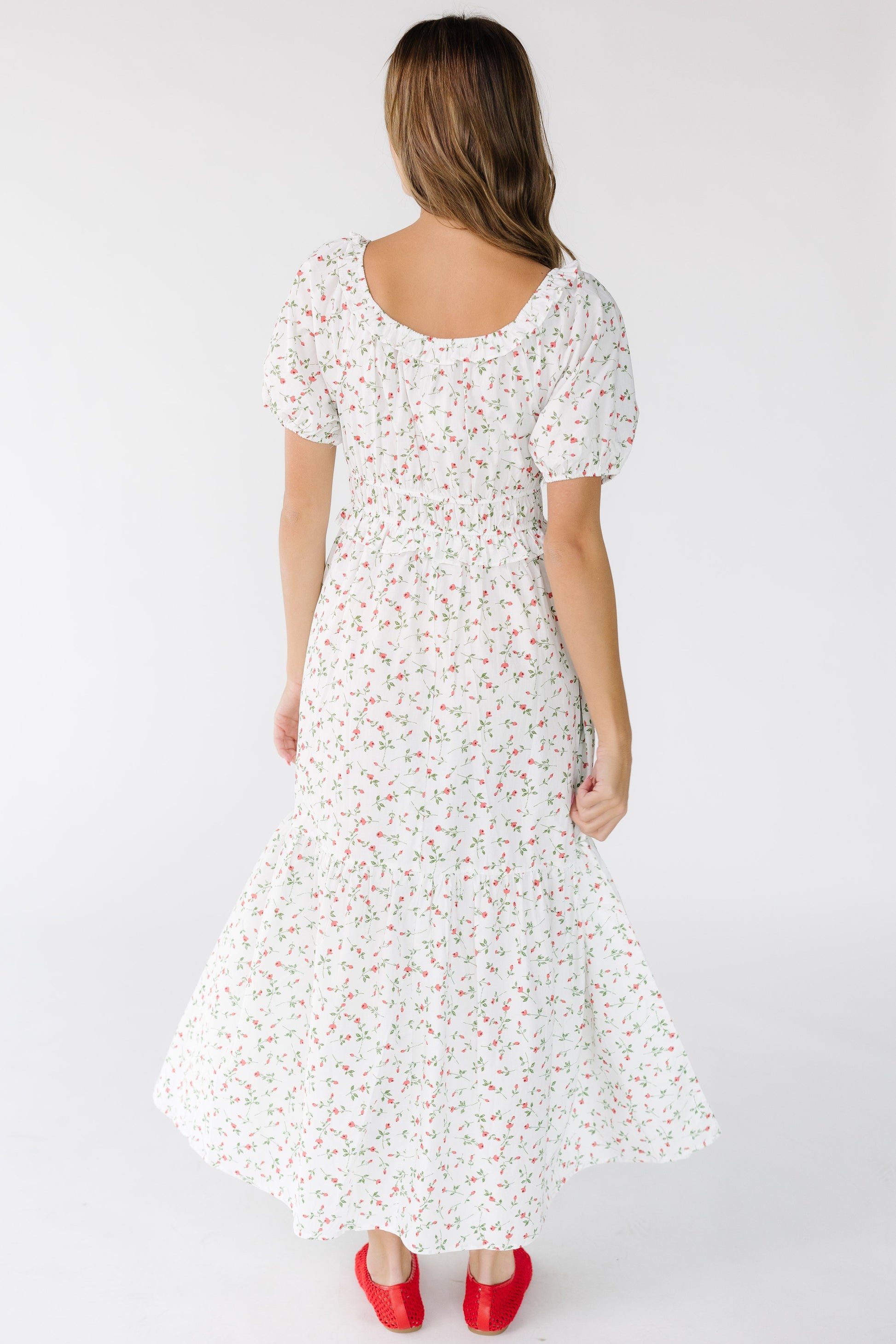 Back view of a young woman wearing an ivory and red floral print dress with a full skirt and short sleeves.