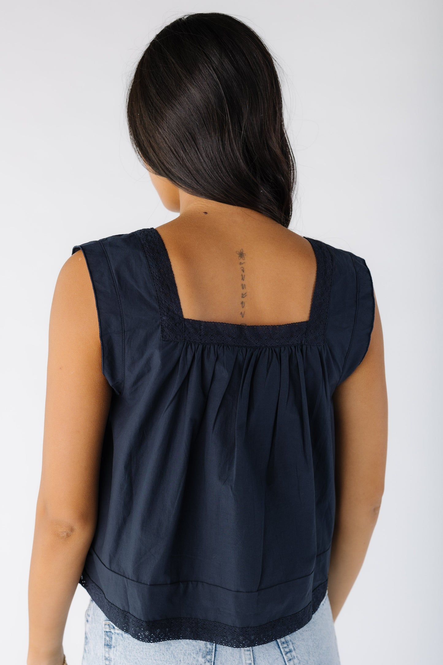 Back view of a young woman wearing a navy loose fitting, sleeveless top with a back square neckline, and lace trim.