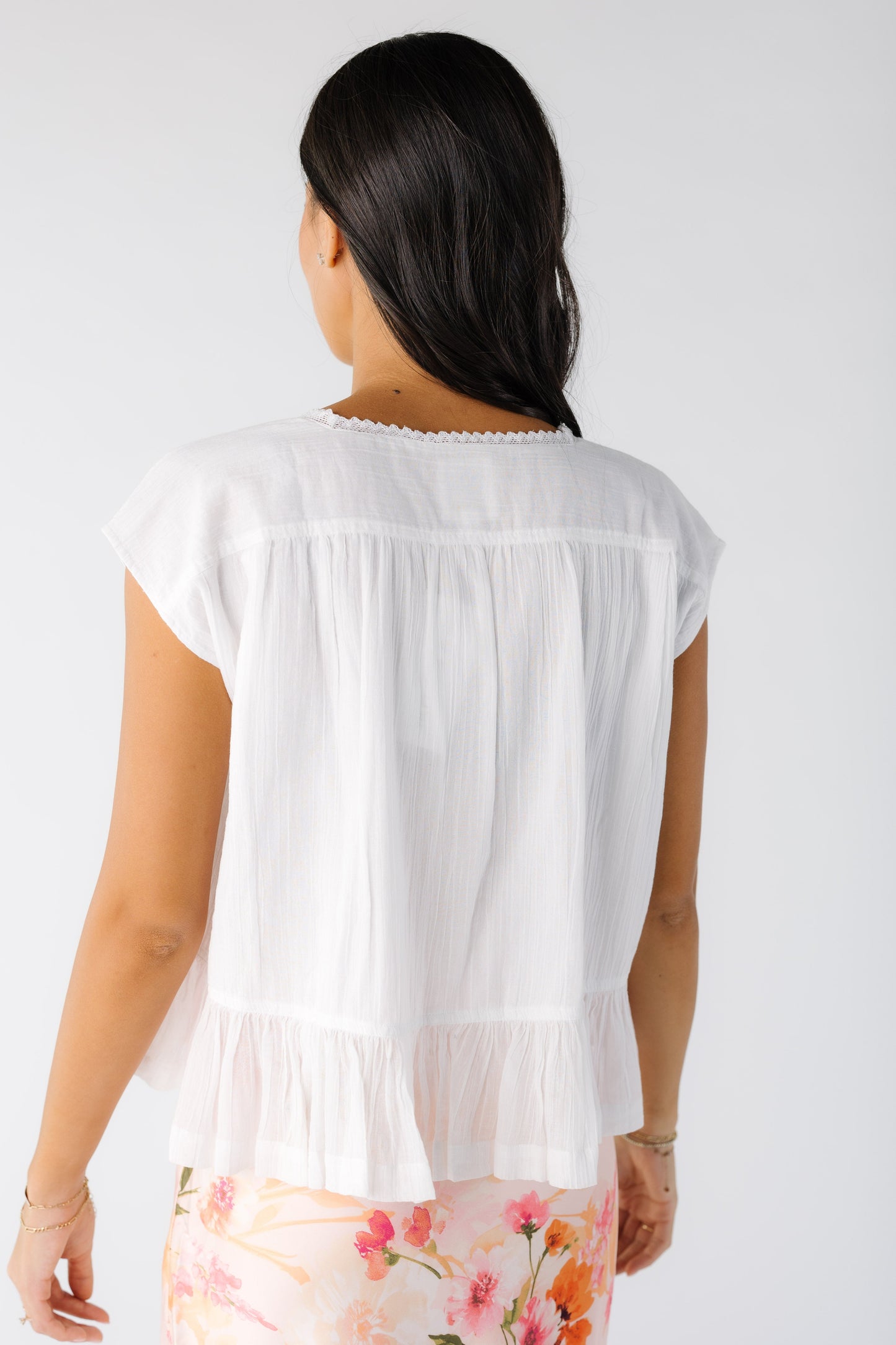 Back view of a young woman wearing a white short sleeve, loose fitting blouse with a bottom ruffle.