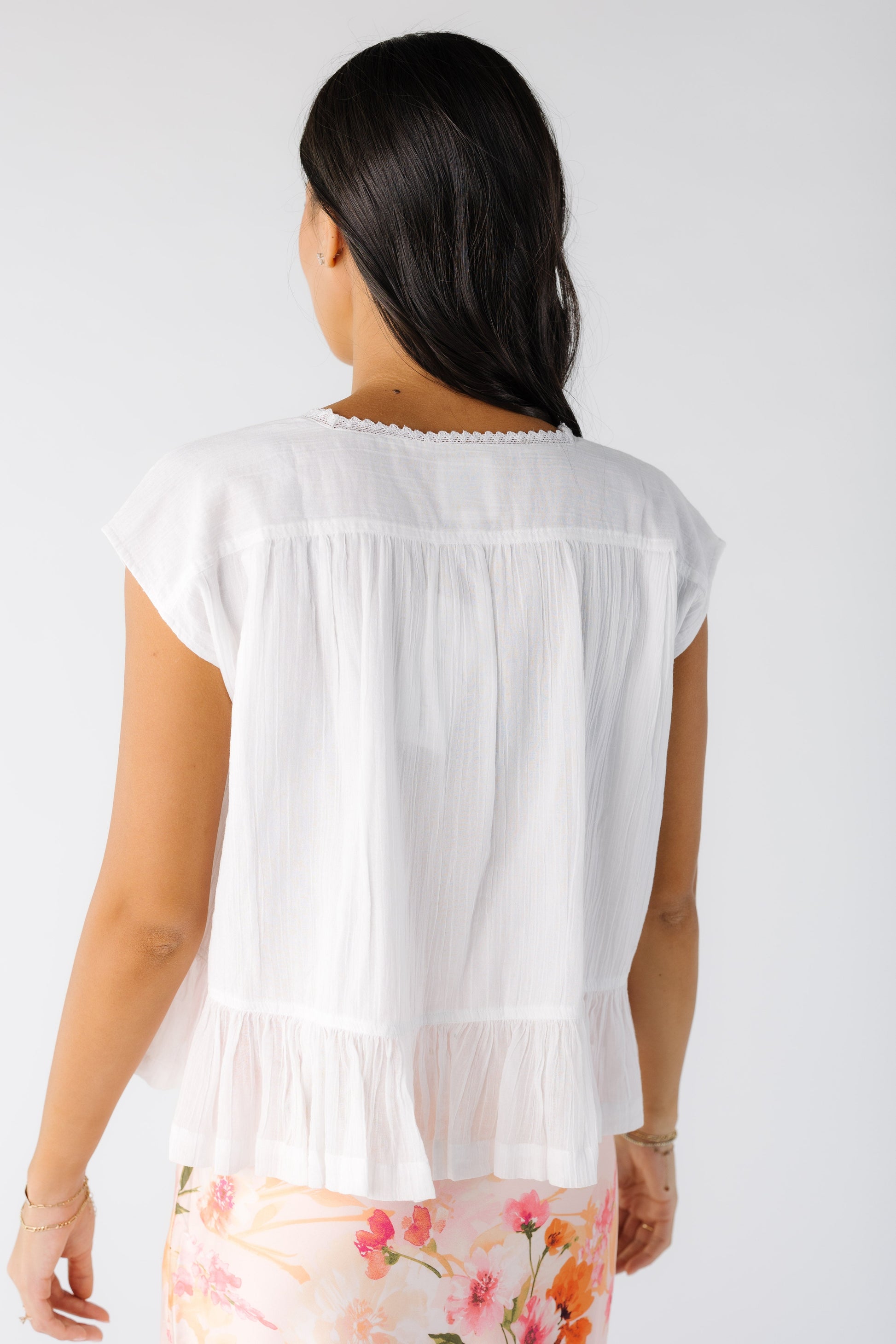 Back view of a young woman wearing a white short sleeve, loose fitting blouse with a bottom ruffle.
