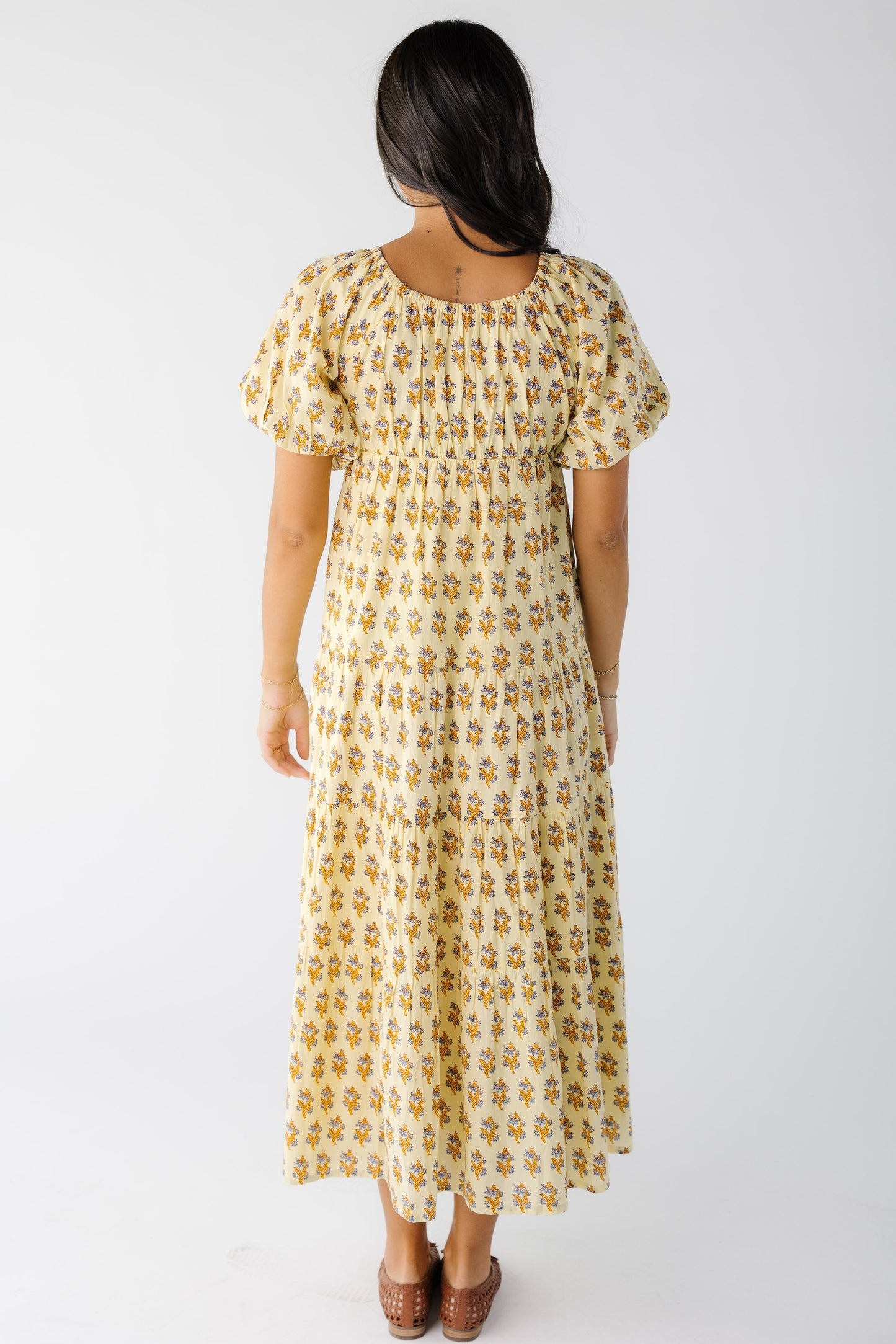 Back view of a woman wearing a yellow floral print midi-maxi dress with a back scoop neckline and large puff sleeves.