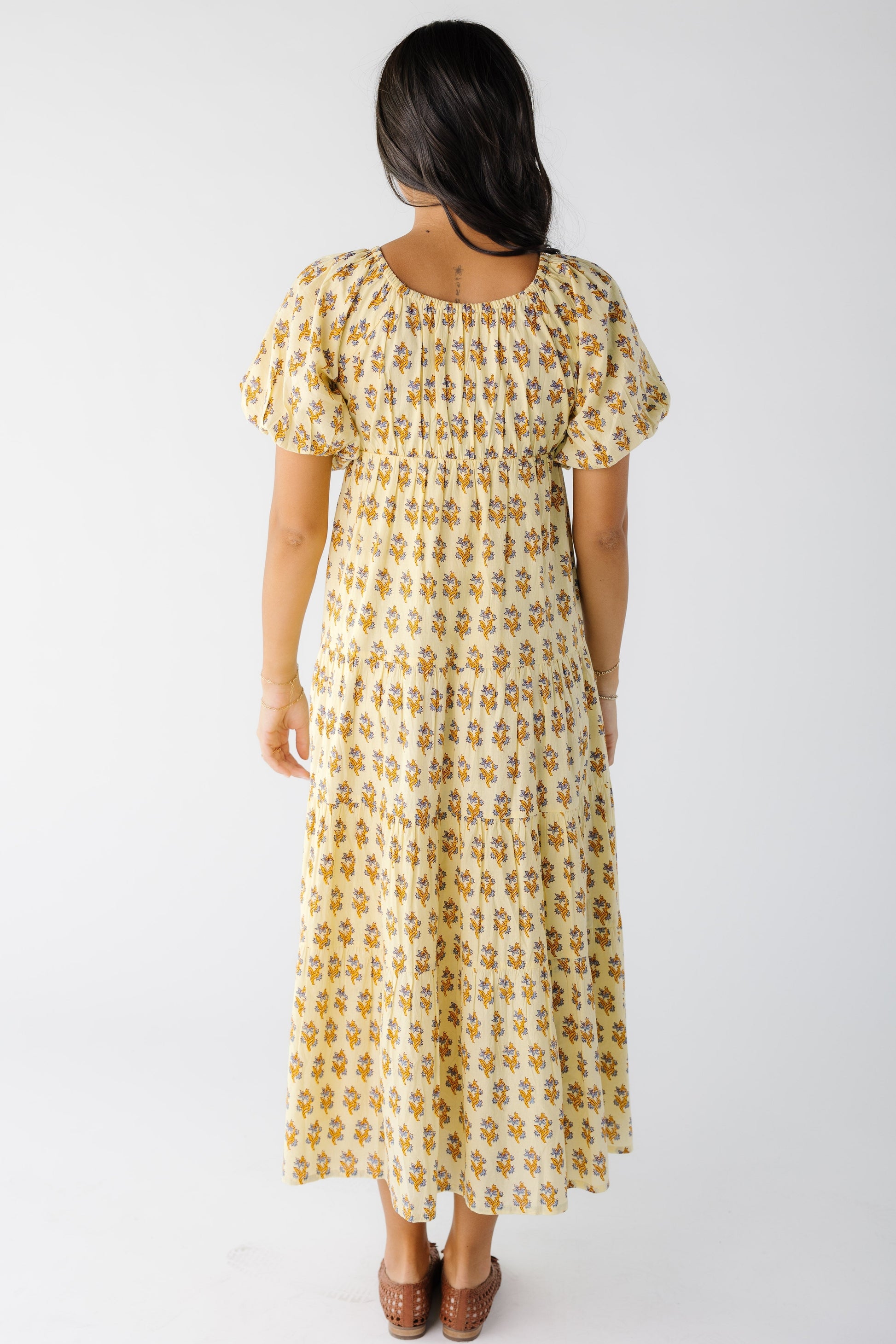 Back view of a woman wearing a yellow floral print midi-maxi dress with a back scoop neckline and large puff sleeves.