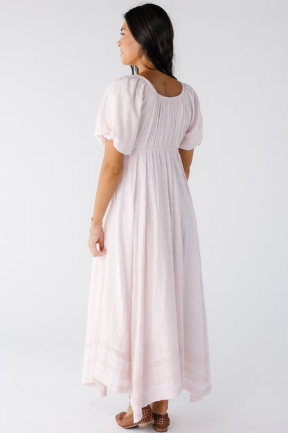 The back view of a wearing a light pink midi-maxi dress with a kerchief hemline with 2 rows of lace insets near the hem.