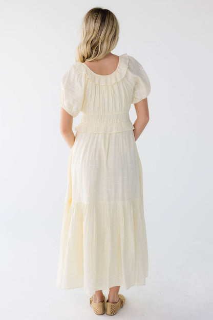 Back view of a woman wearing a light yellow midi-maxi dress with a scoop, ruffled neckline and short puff sleeves.