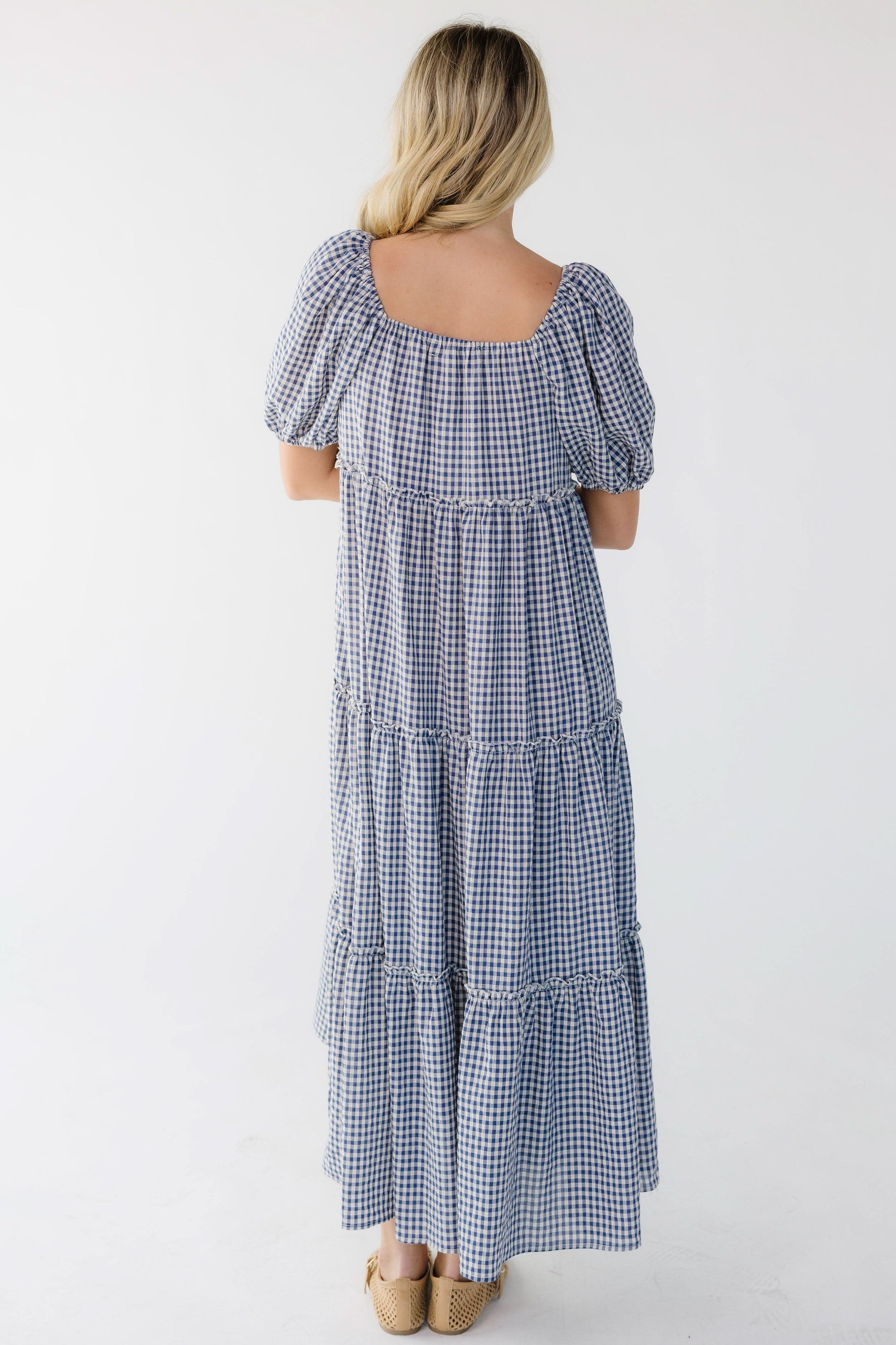Back view of a woman wearing a blue gingham tiered dress with balloon sleeves and a square neckline.