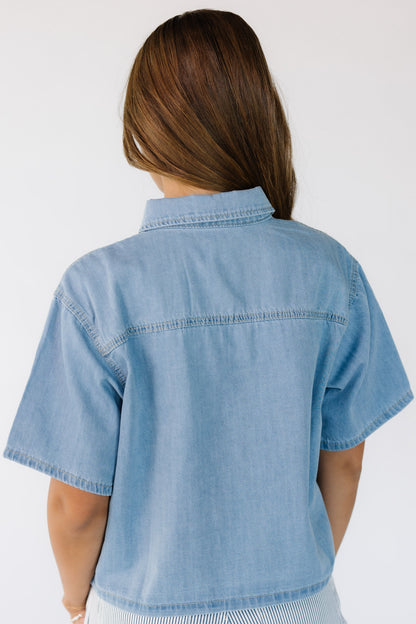 Back view of a young woman wearing a light denim shirt with short sleeves and straight cut hem.