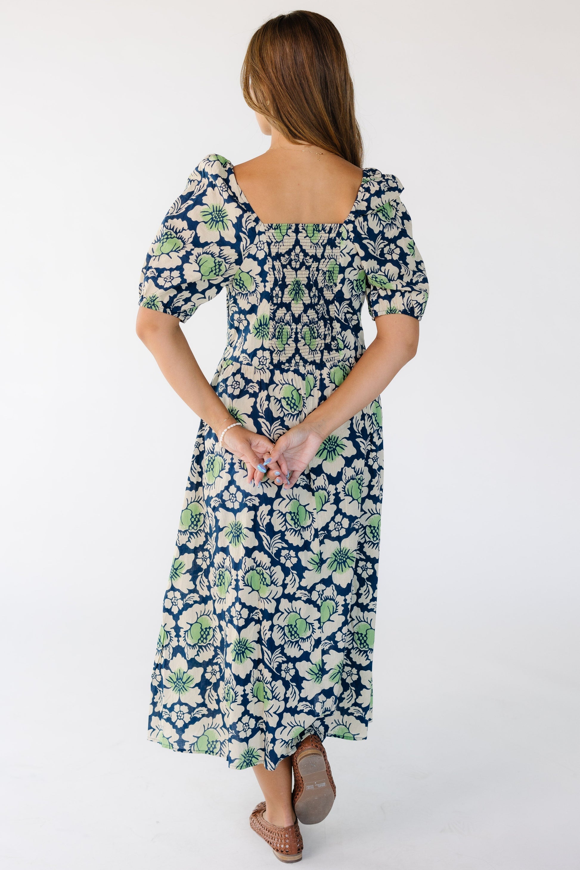 Back view of a young woman wearing a blue and green floral print midi dress with a elastic smocked back bodice panel, a square neckline with large balloon sleeve.
