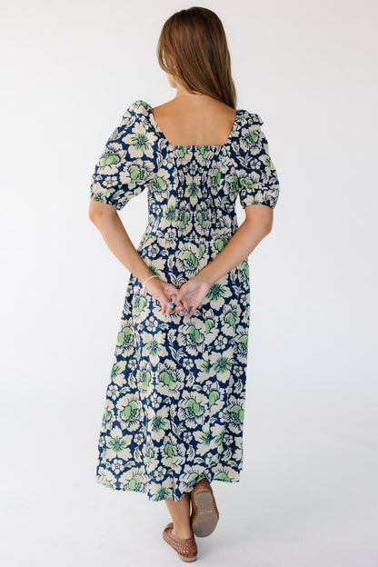 Back view of a young woman wearing a blue and green floral print midi dress with a elastic smocked back bodice panel, a square neckline with large balloon sleeve.