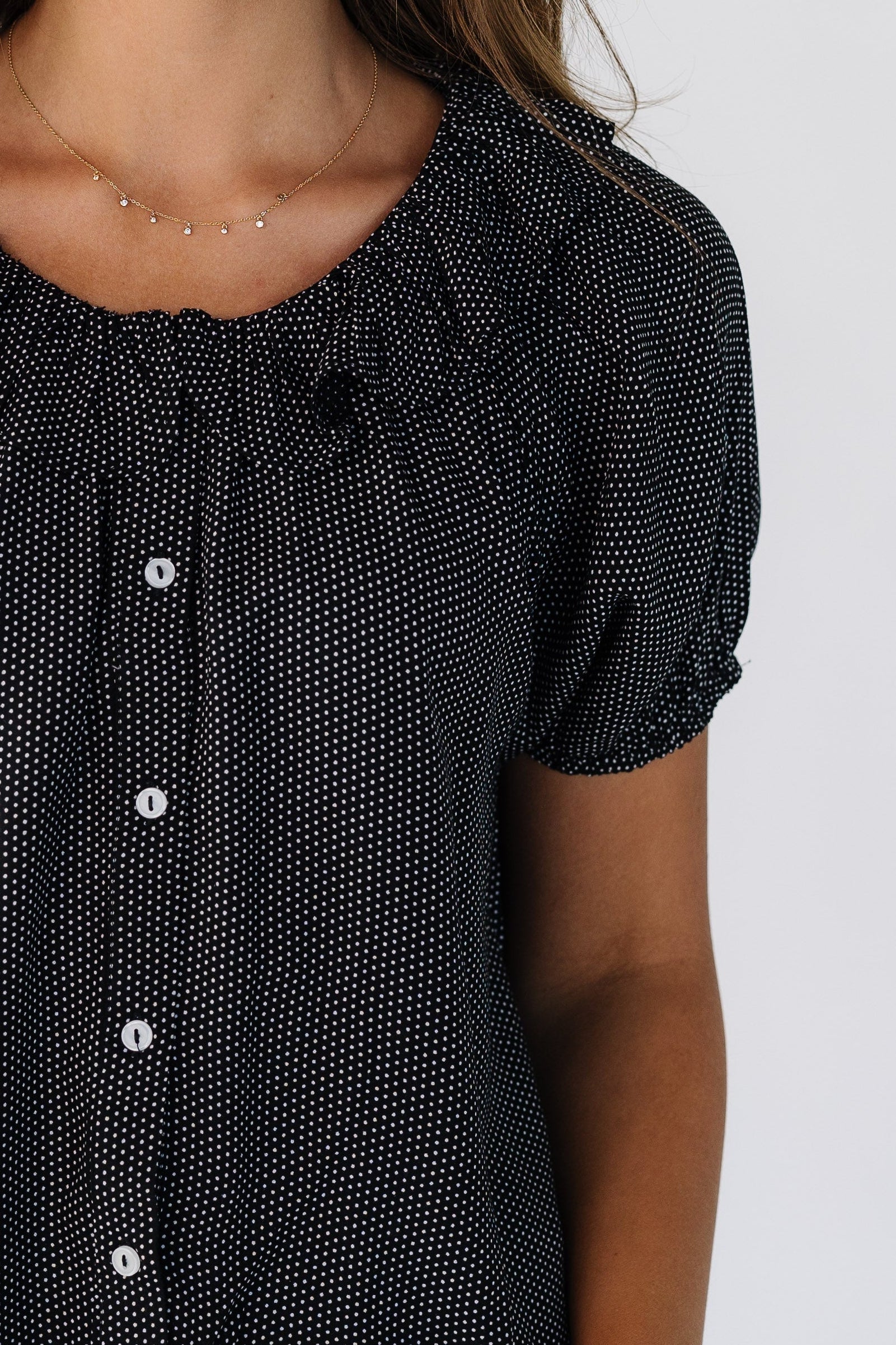 Close view of a woman wearing a black & white polka dot button down top with short puff sleeves and a round ruffled collar.