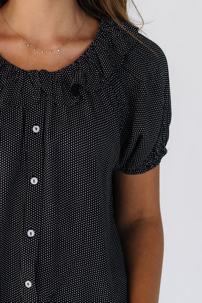 Close view of a woman wearing a black & white polka dot button down top with short puff sleeves and a round ruffled collar.