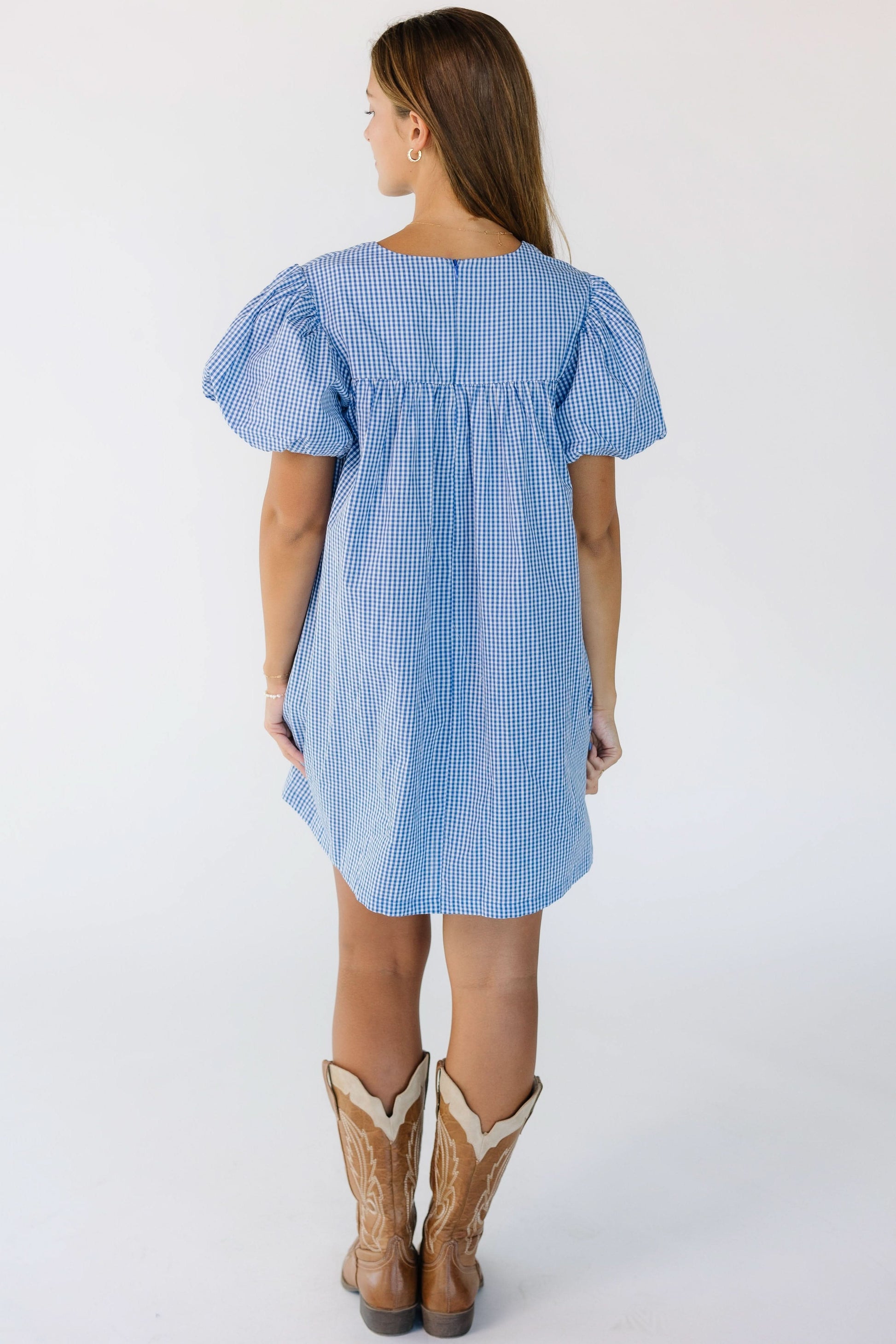 Back view of a young woman wearing a blue and white gingham short dress with short puff sleeves.
