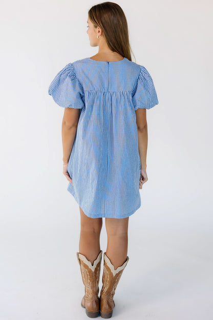 Back view of a young woman wearing a blue and white gingham short dress with short puff sleeves.