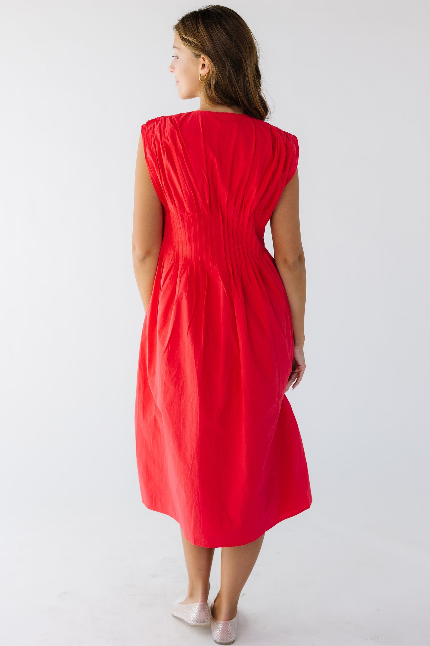 Back view of a woman wearing a red sleeveless midi dress with a closer fitting waist and pleats at the shoulder.