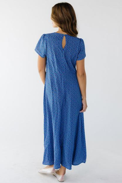Back view of a woman wearing a blue floral midi dress with a back zipper, keyhole & button closure and short sleeves.
