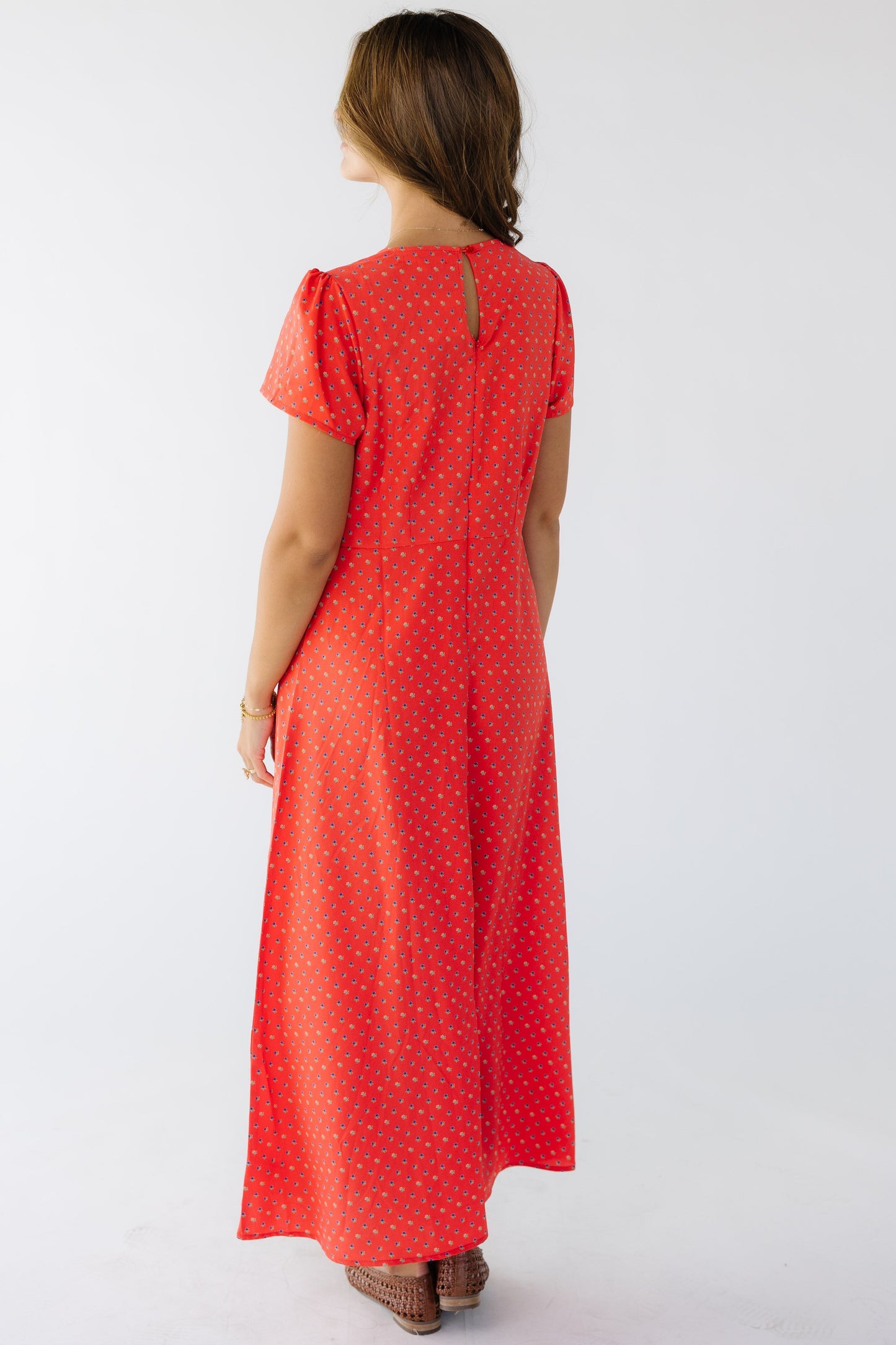 Back view of a young woman wearing a red floral midi sheath dress with a back zipper closure with a button and keyhole closure and short sleeves.