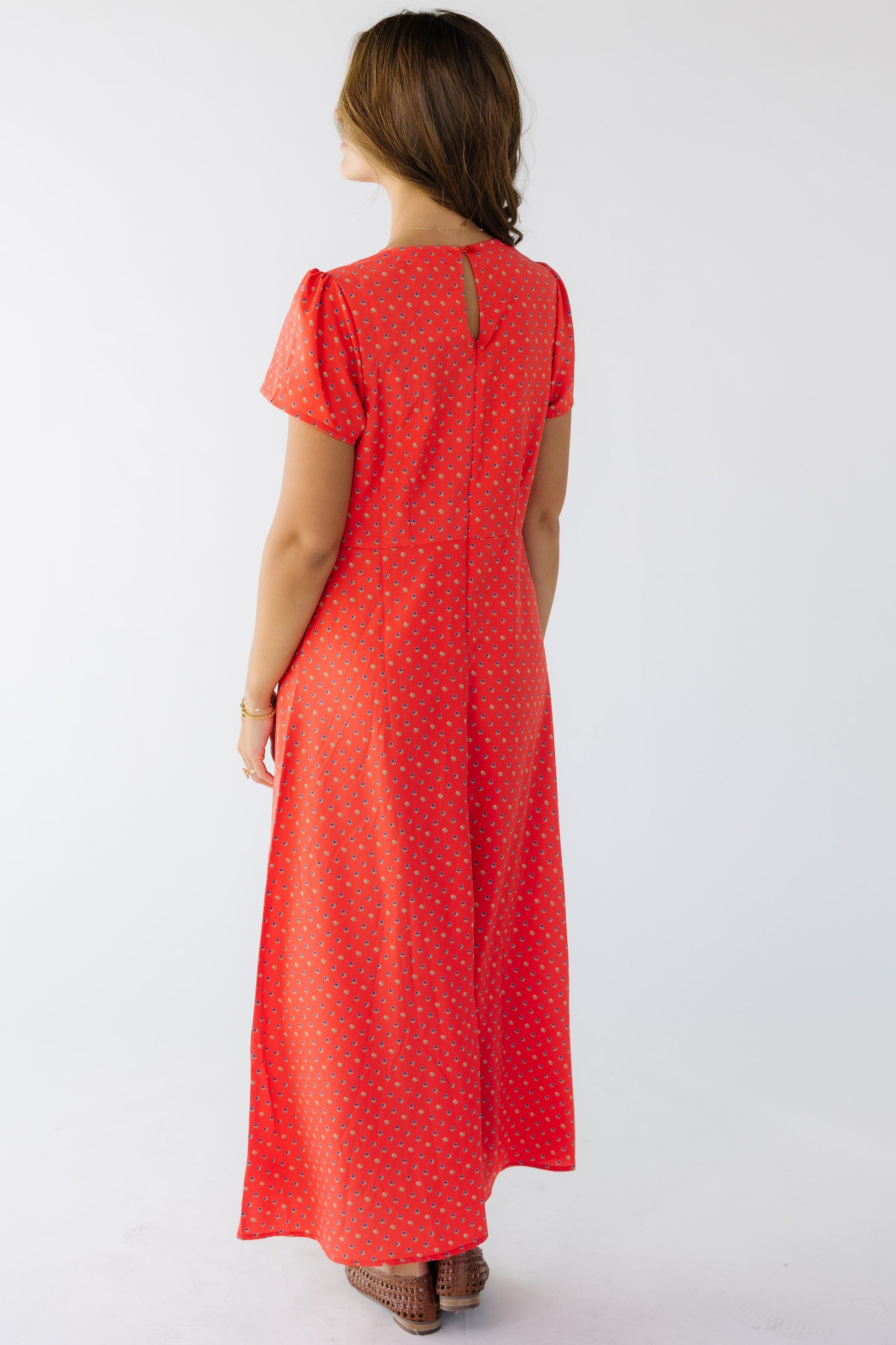 Back view of a young woman wearing a red floral midi sheath dress with a back zipper closure with a button and keyhole closure and short sleeves.