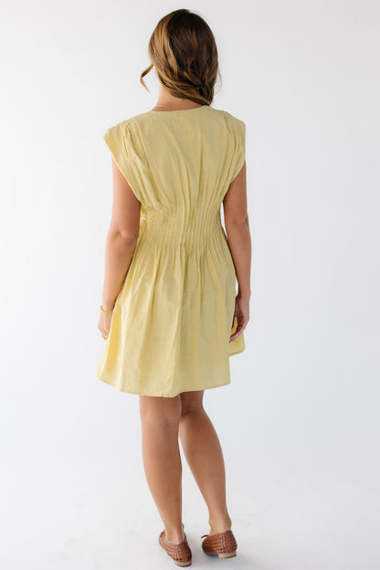 Back view of a woman wearing a yellow sleeveless short dress with pleats all around the waist.