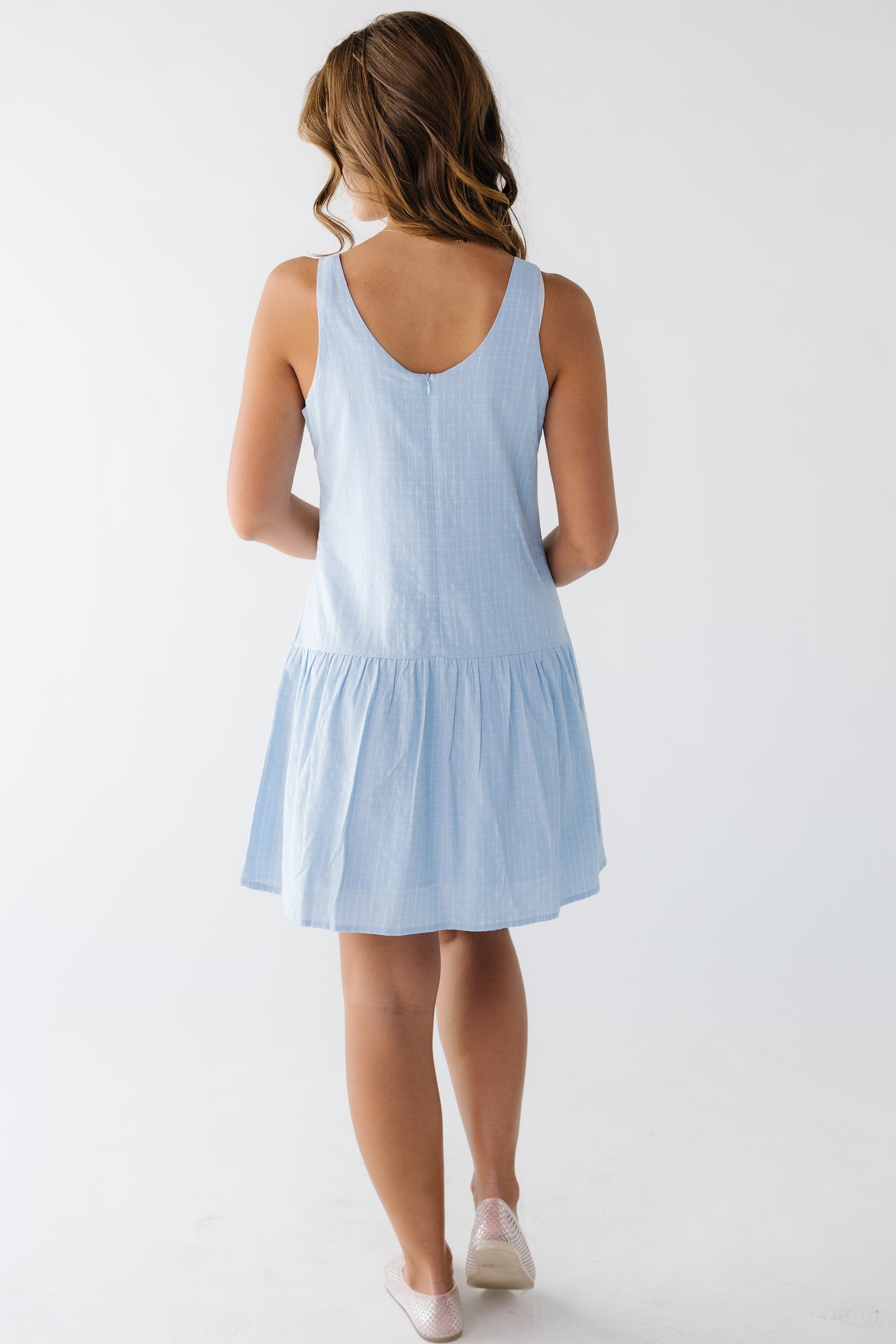 Back view of a young woman wearing a sleeveless blue plaid short dress with a back scoop neckline and drop waist.