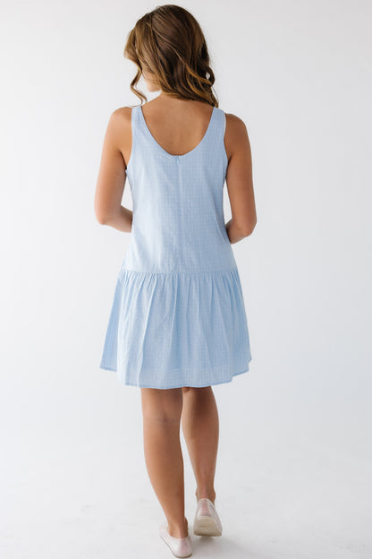 Back view of a young woman wearing a sleeveless blue plaid short dress with a back scoop neckline and drop waist.