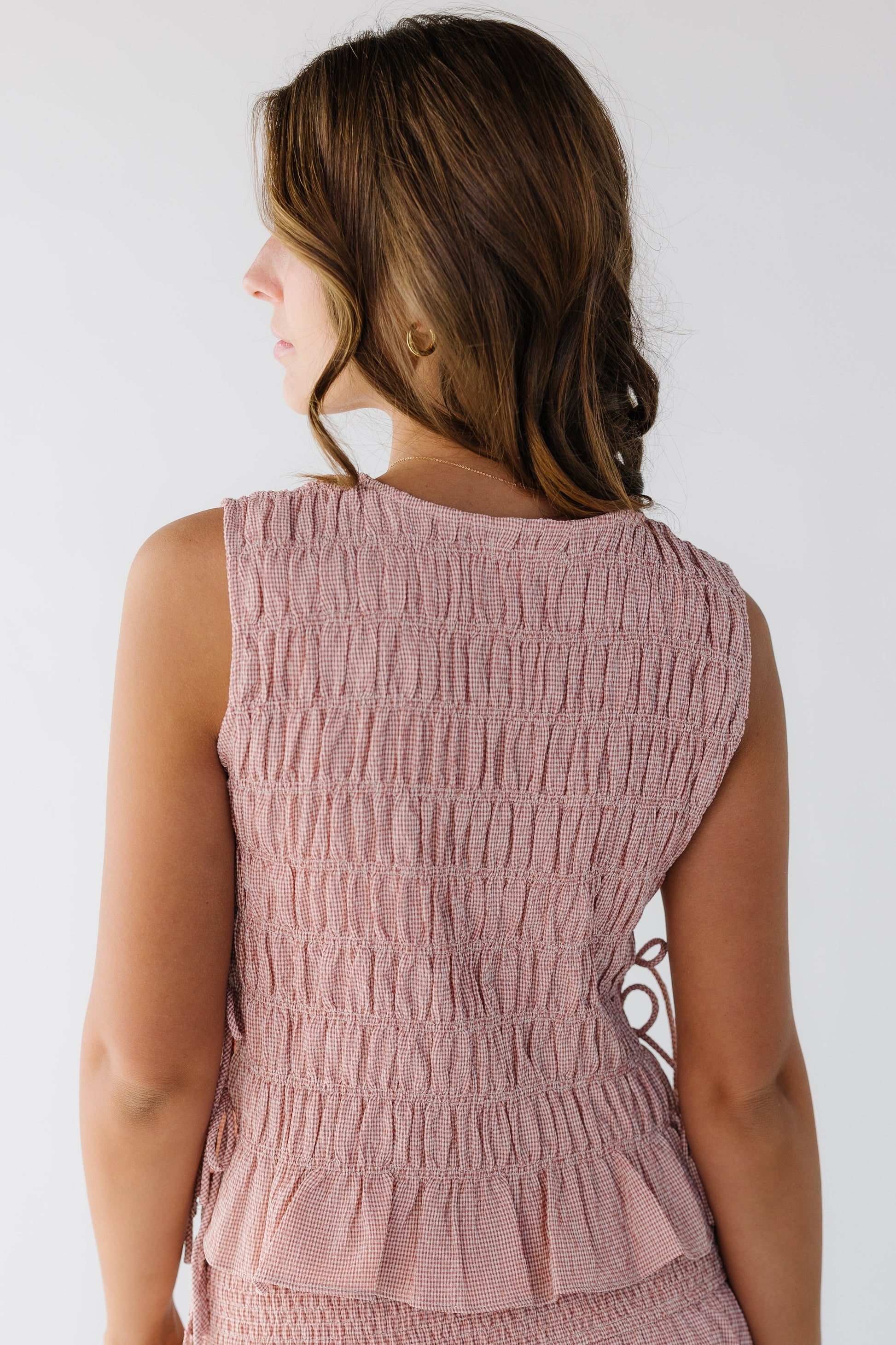 Back view of a young woman wearing a pink gingham sleeveless smocked blouse.