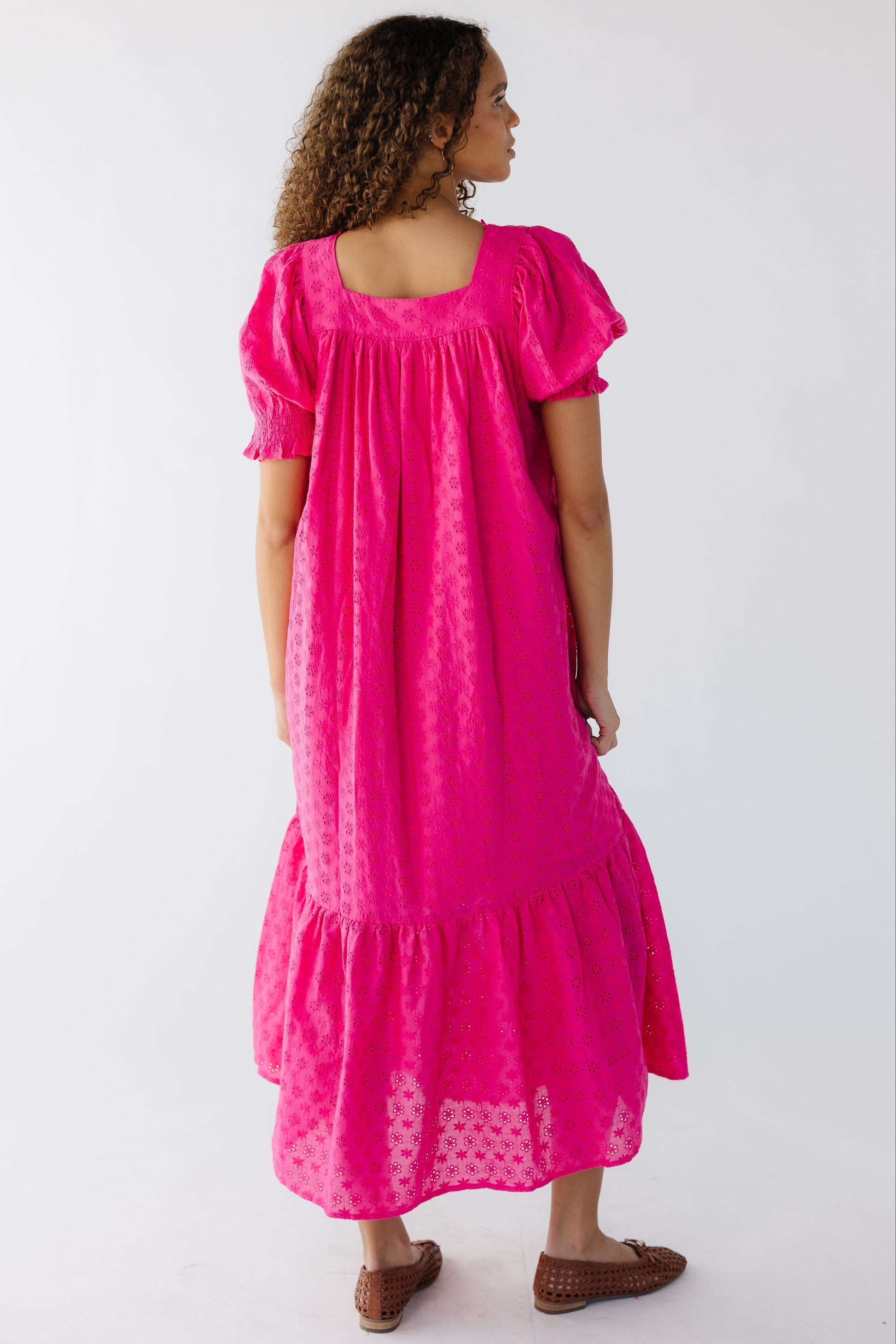 Back view of a teenage girl wearing a bright pink eyelet midi dress with a square back neckline and large puff sleeves with elastic smocked cuff.