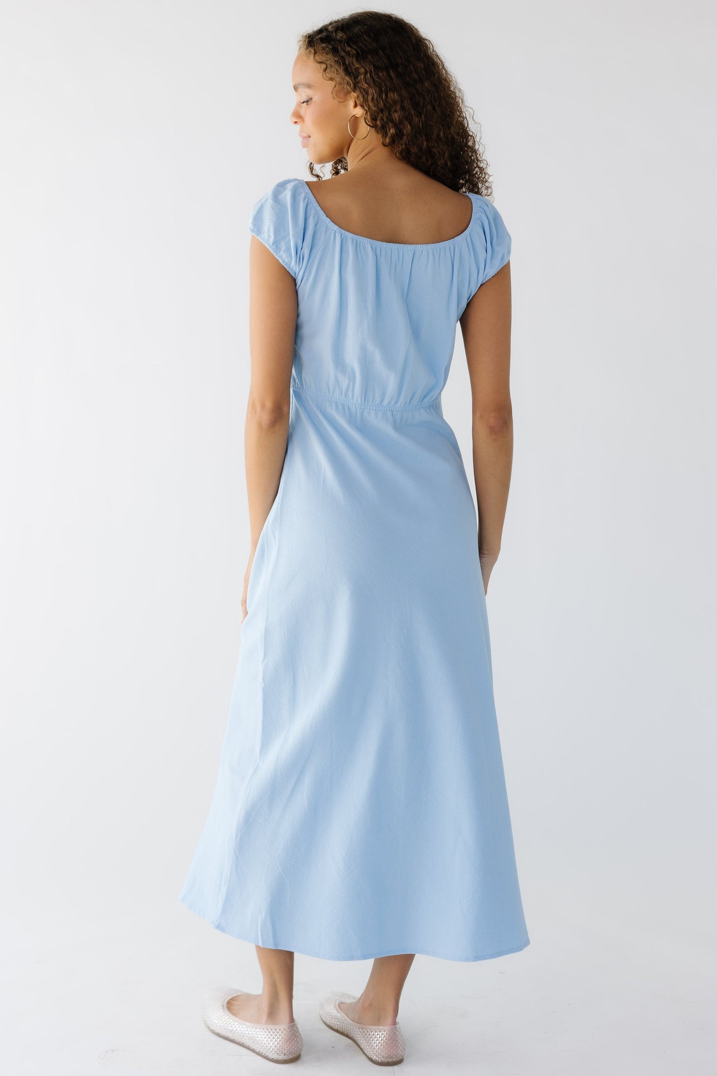 Back view of a teen girl wearing a close fitting light blue midi dress with a scoop neckline, elastic back waist and short sleeves.