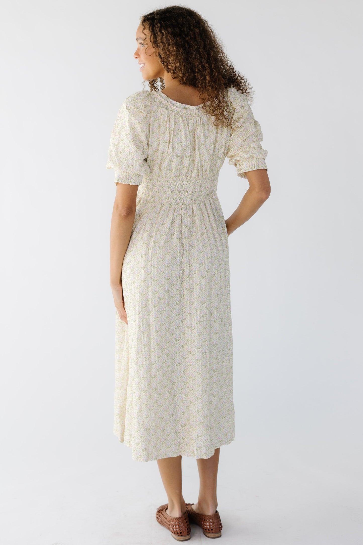 Back view of a young woman wearing a cream floral midi dress with short balloon sleeves and a wide elastic waist.