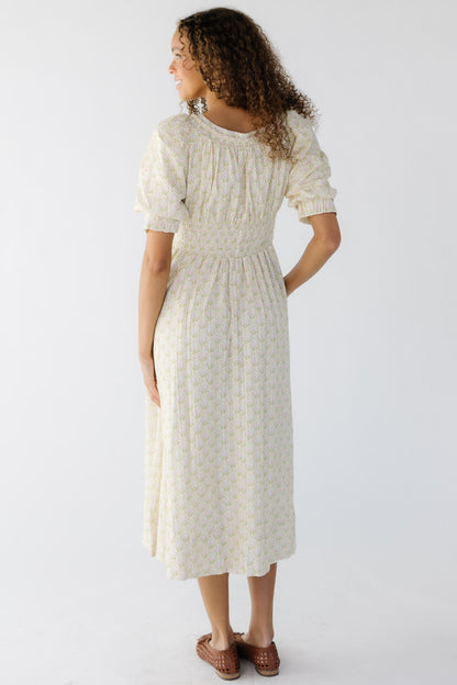 Back view of a young woman wearing a cream floral midi dress with short balloon sleeves and a wide elastic waist.