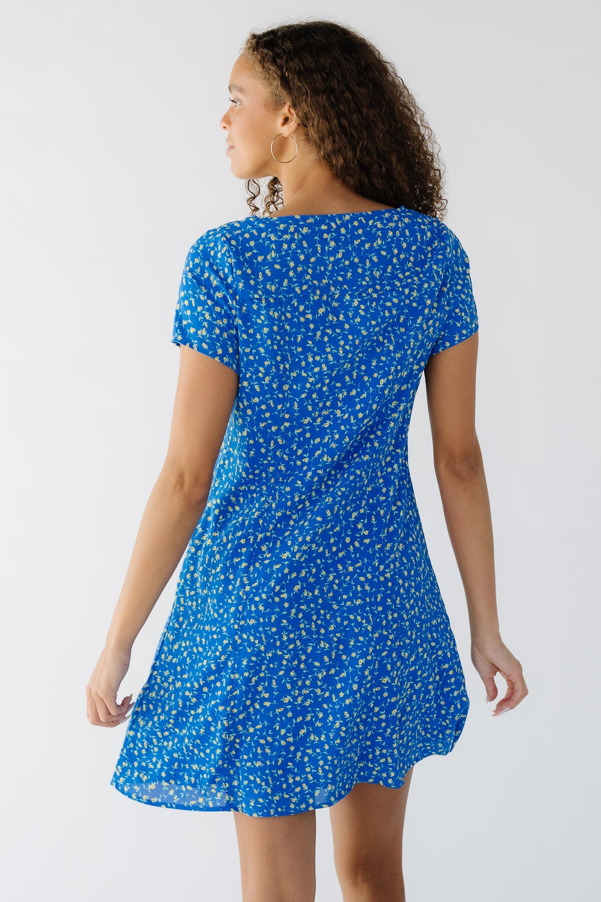 Back view of a teen girl wearing a blue floral dress cut above the knee with short sleeves.