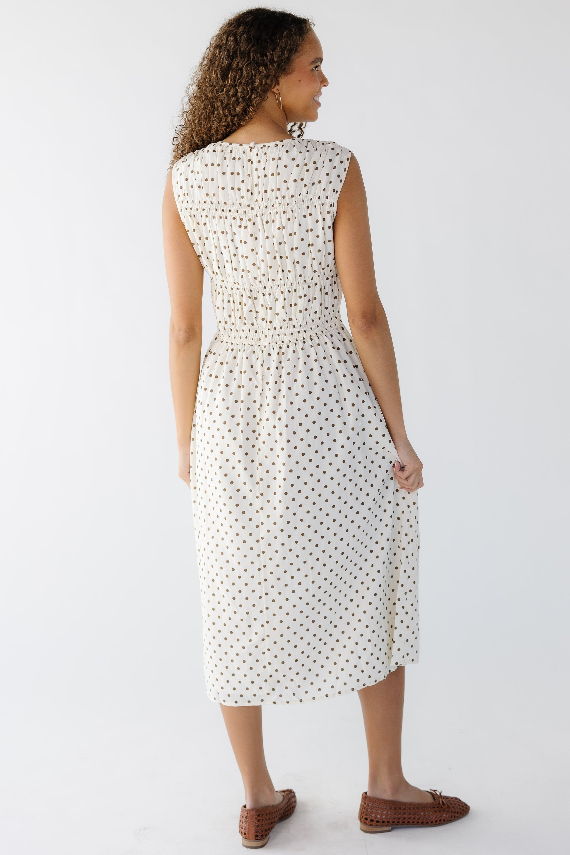 Back view of a young woman wearing a cream and brown polka dot midi, sleeveless dress.
