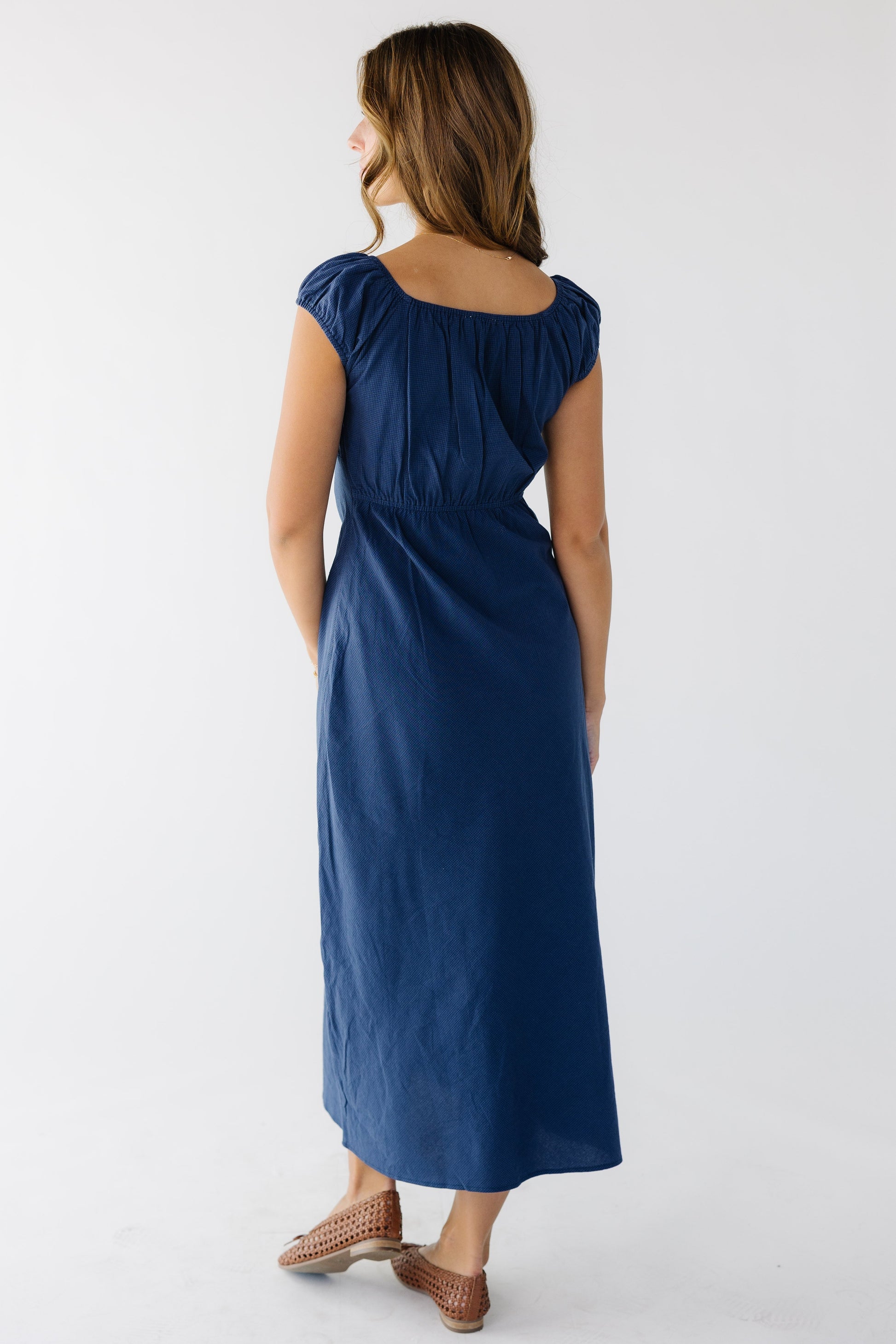 Back view of a woman wearing a navy A-line midi dress with an elastic back waist with a scoop neckline and short puff sleeves.