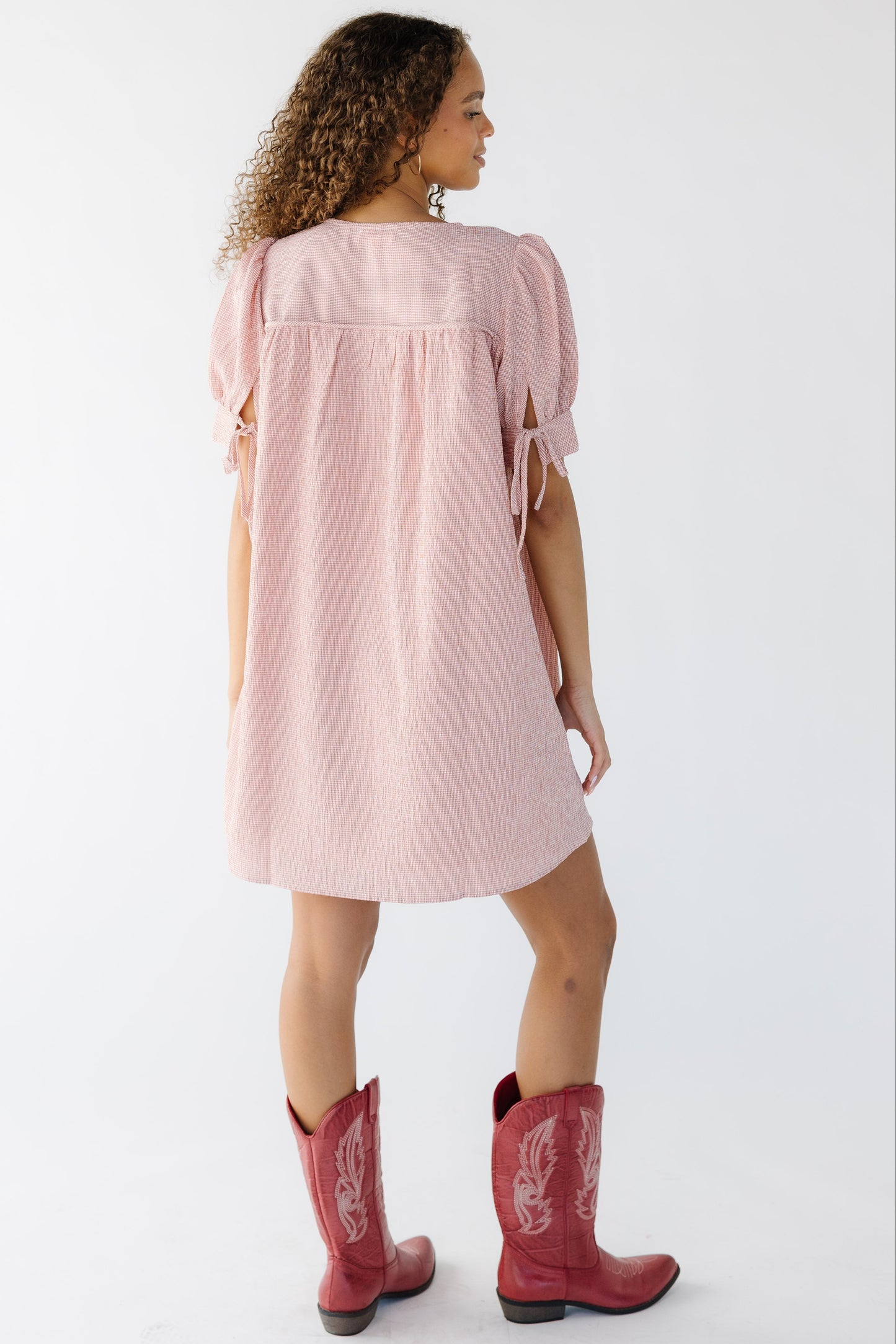 Back view of a woman wearing a pink gingham short dress with short puff sleeves.