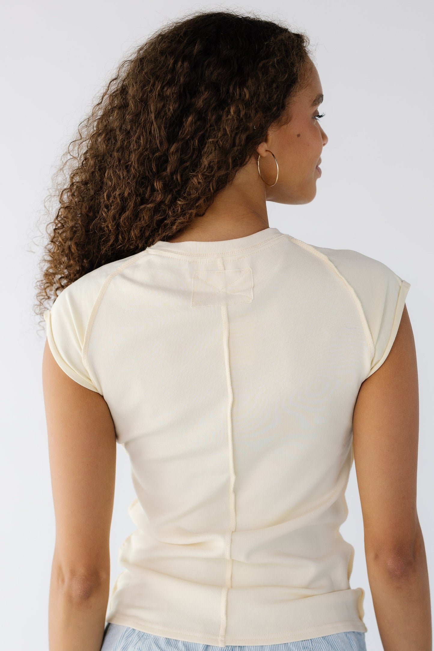 Back view of a teen girl wearing a light yellow rib knit top with exposed seams and smooth fabric sleeves.
