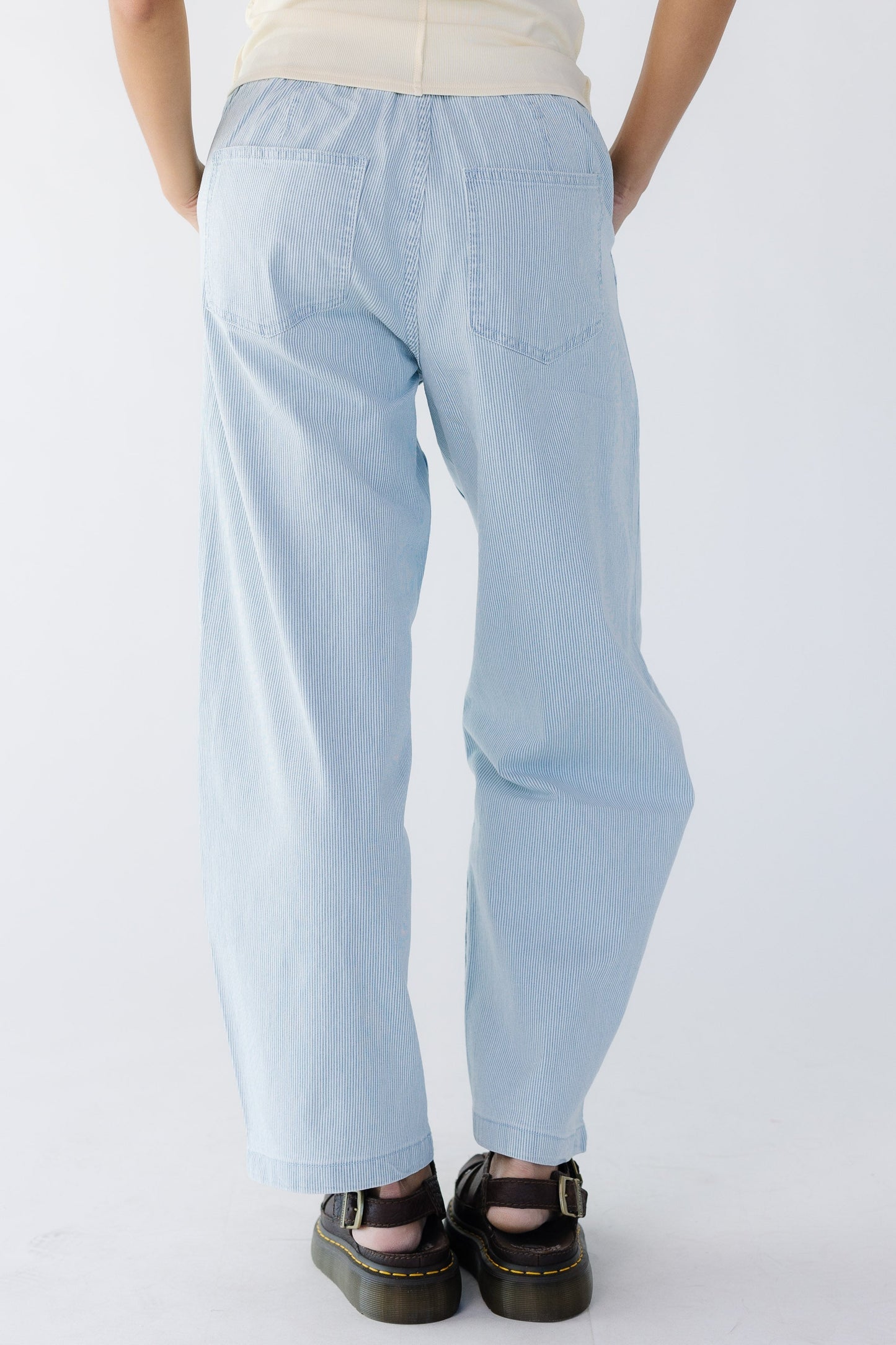 Back view of a young woman wearing a pair of blue & white pinstripe denim wide leg pants with back pockets.