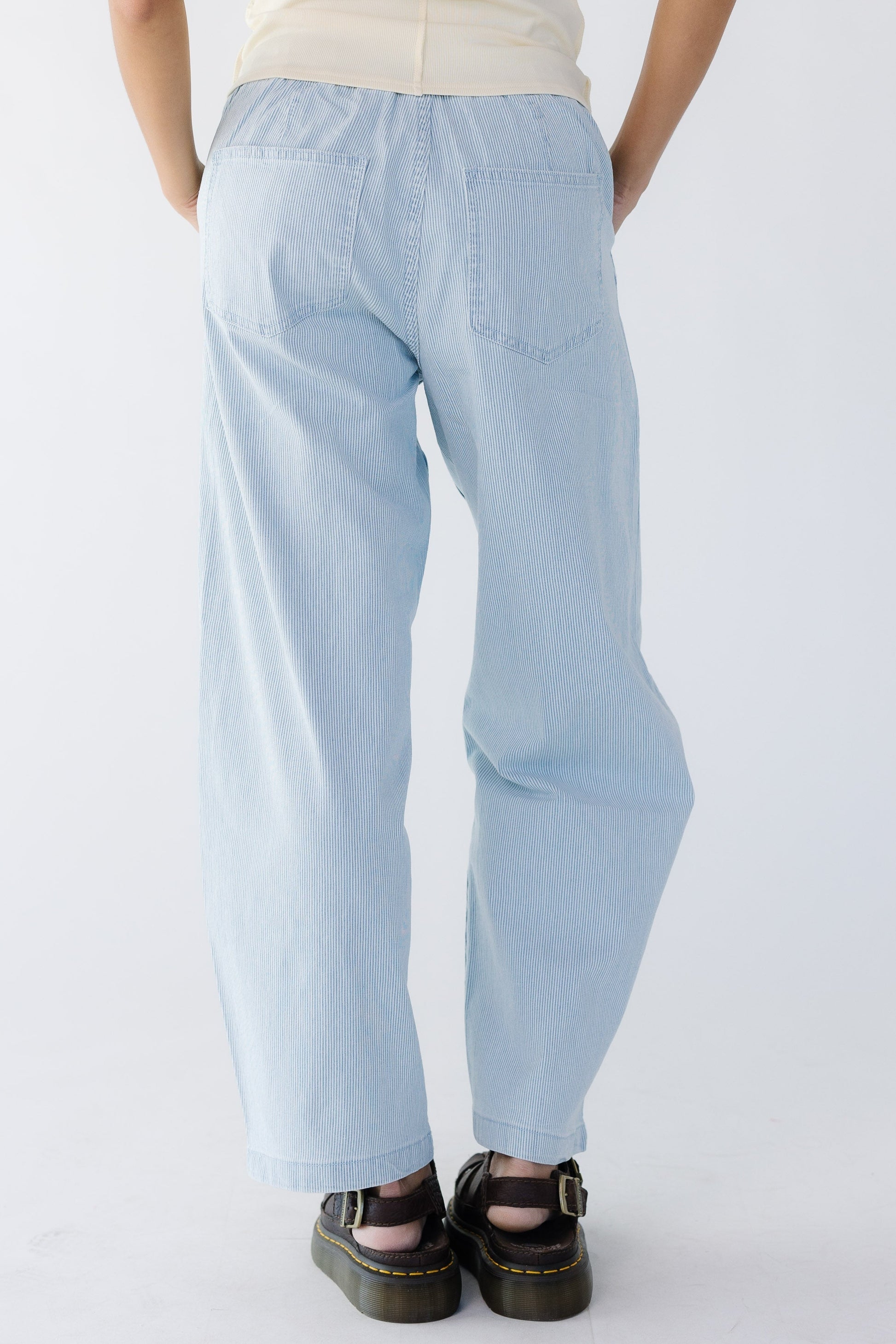 Back view of a young woman wearing a pair of blue & white pinstripe denim wide leg pants with back pockets.