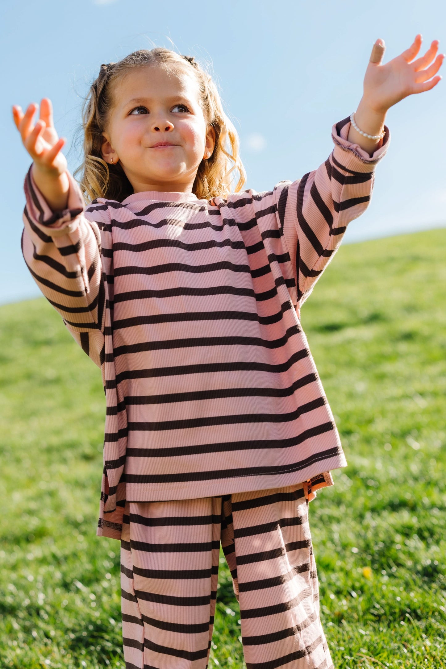 Little girl wearing a modest pink and brown stripe lounge set with a crew neckline, long sleeves and wide leg pants.