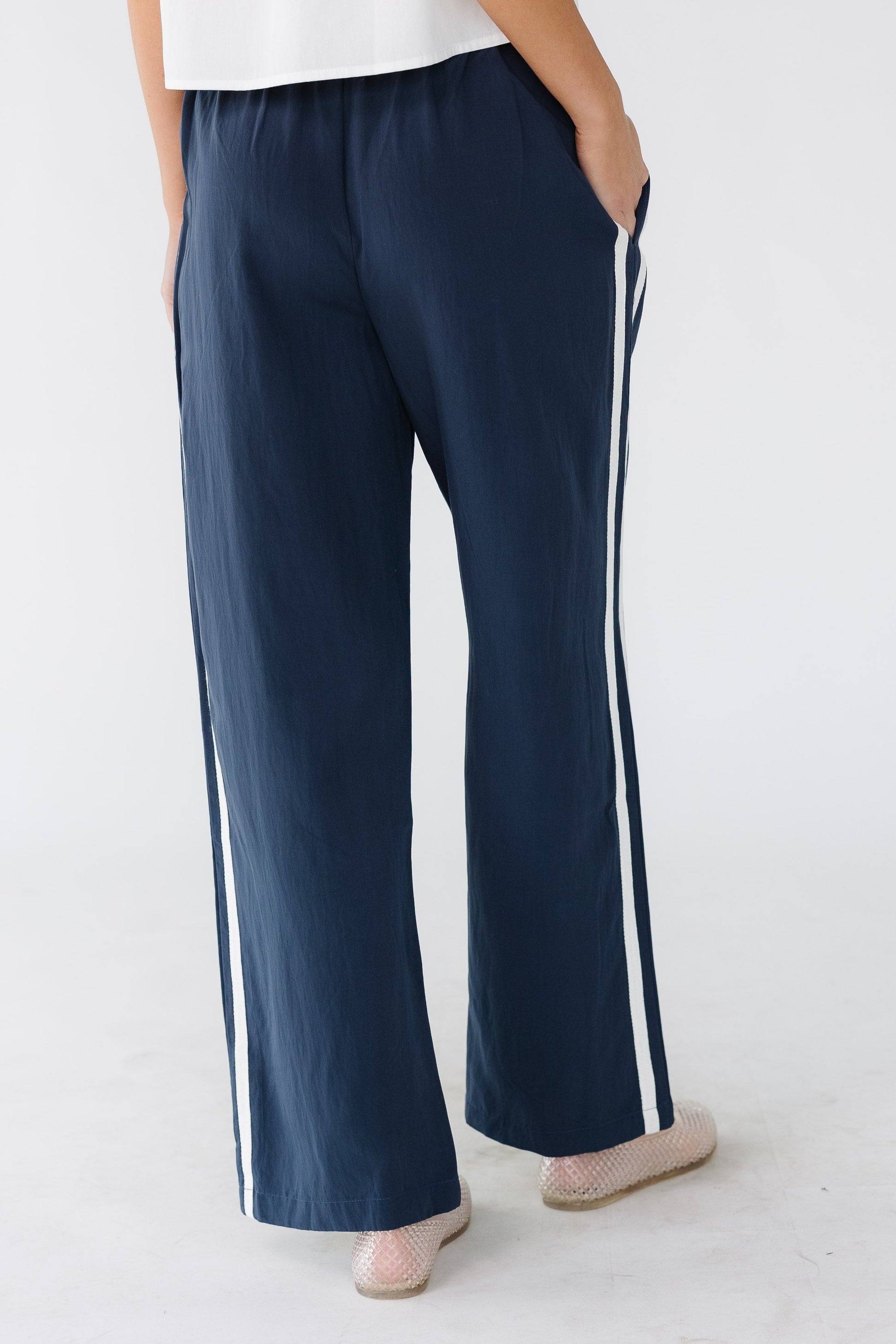 Back view of a woman wearing a pair of navy wide leg track pant with side pockets and an elastic waist.