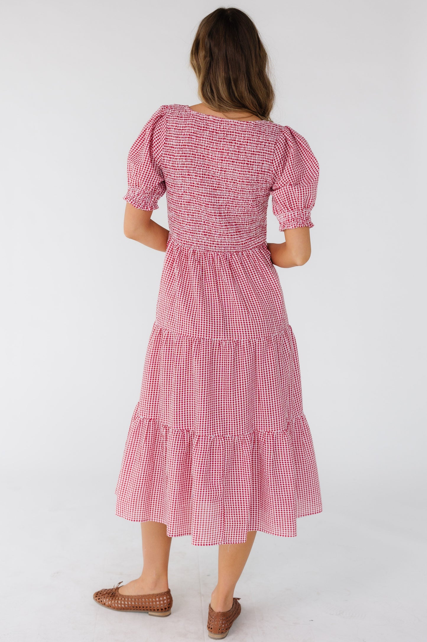 Back view of a young woman wearing a red & white gingham midi dress with multiple gathered tiers, smocked bodice and short puff sleeves.