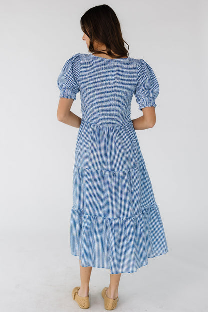 Back view of a young woman wearing a blue & white gingham midi dress with a smocked bodice and short puff sleeves.