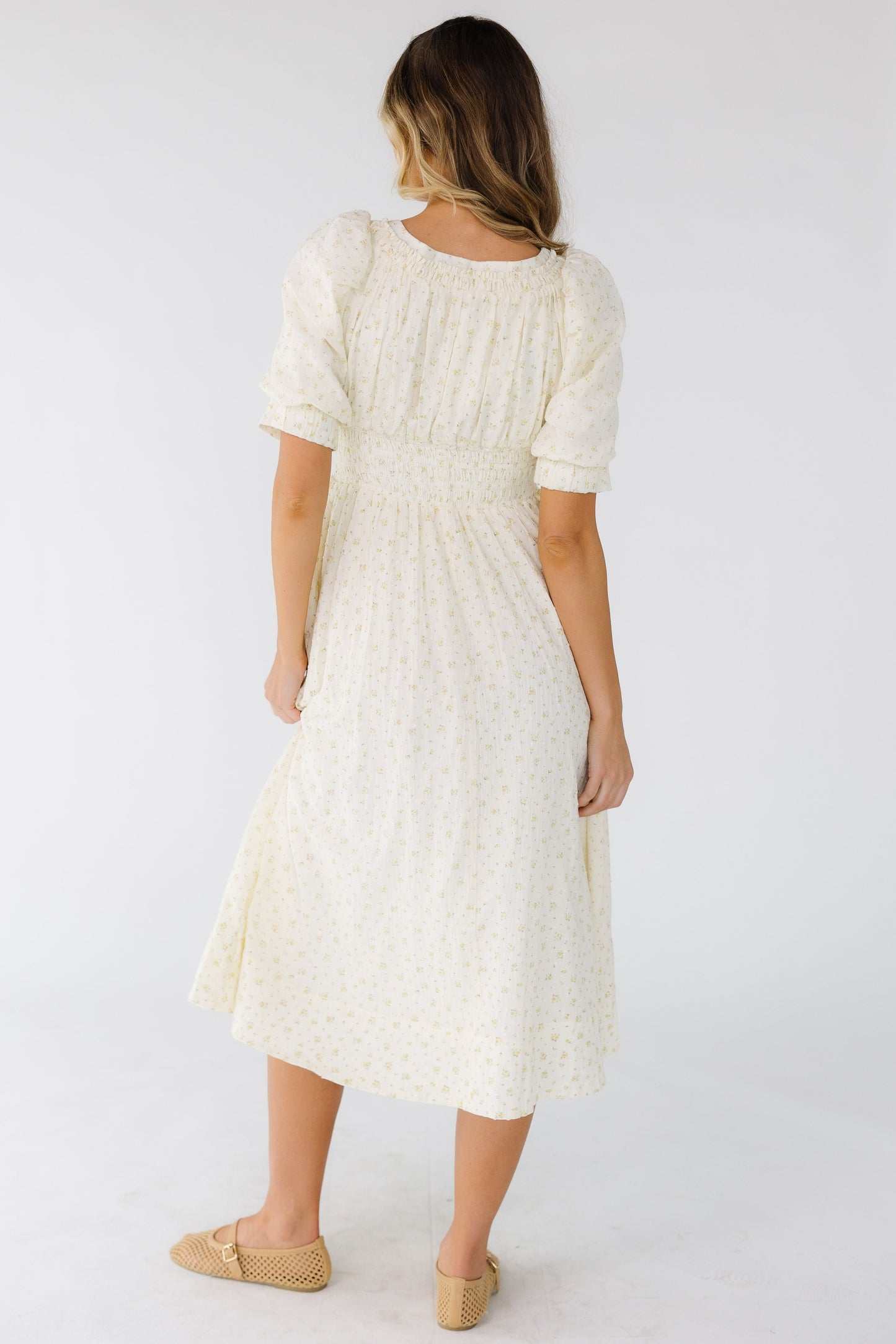 Back view of a woman wearing a cream floral midi dress with s wide waistband and balloon sleeves.